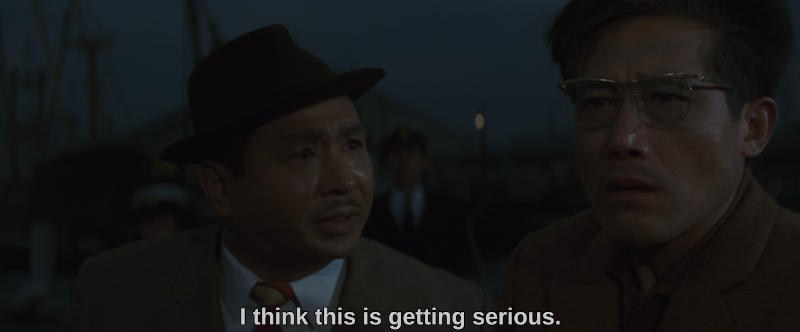 Color image. Two men stand at a dockyard, one looking off camera and the other speaking to him. The subtitle reads, I think this is getting serious.