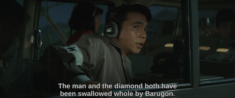 Color image. A radio announcer with a frightened face speaks into his receiver. The subtitle reads, The man and the daimond both have been swallowed whole by Barugon.