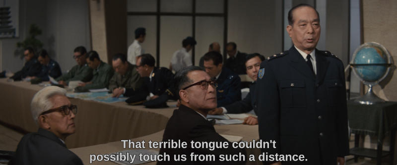 Government and military officials sit at a long table and look off screen. One of them says, That terrible tongue couldn't possibly touch us from such a distance.