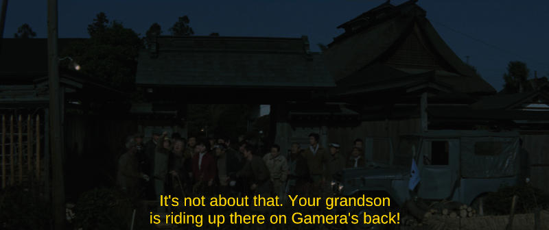 A group of people gather in front of a large traditional Japanese rural home, looking towards the sky. The subtitle reads, It's not about that. Your grandson is riding up there on Gamera's back!