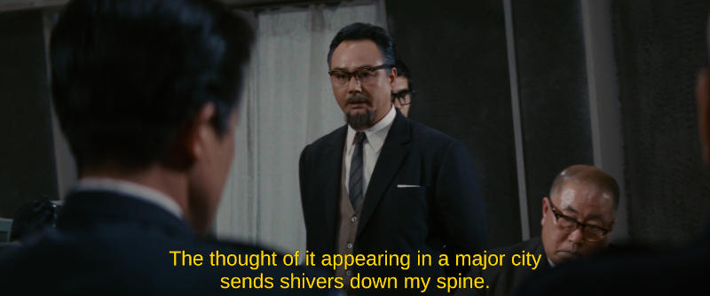 A pensive scientist in a dark suit with a goatee addresses a group of officials. The subtitle reads, The thought of it appearing in a major city sends shivers down my spine.