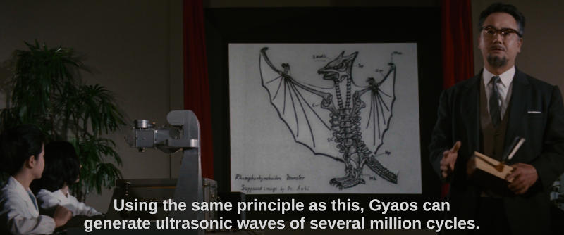 A scientist stands in front of a projected image of a flying dinosaur and holds a tuning fork. The subtitle reads, Using the same principle as this, Gyaos can generate ultrasonic waves of several million cycles.