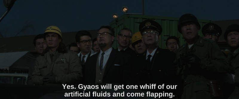 A group of scientists, military men and construction workers stand outside at night, looking into the distance. The subtitle reads, Yes. Gyaos will get one whiff of our artificial fluids and come flapping.